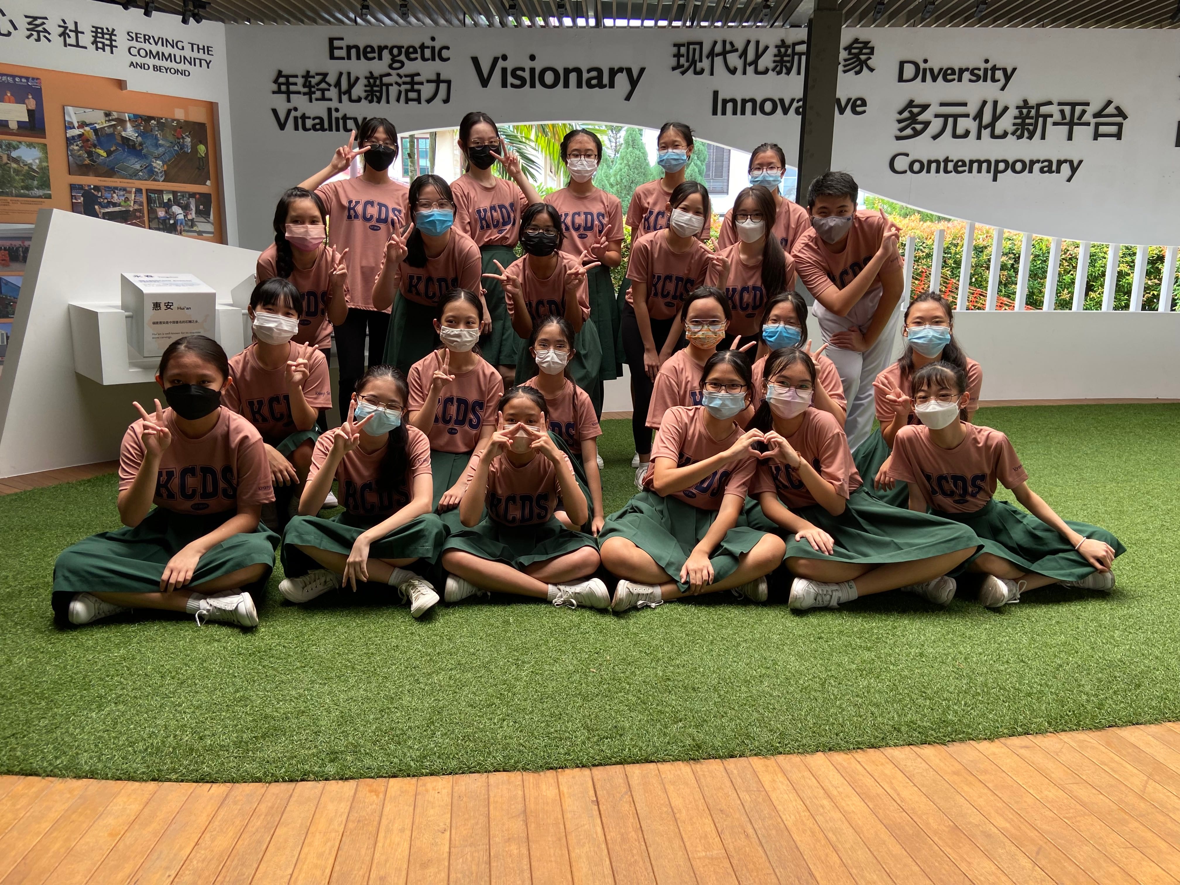 Group of masked students in pink KCDS shirts and green skirts posing with peace signs indoors.